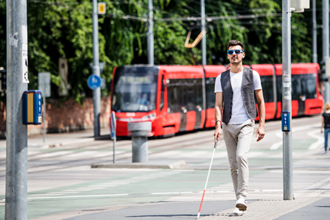 Ein junger blinder Mann läuft auf dem Gehweg. Er hat eine Sonnenbrille auf und einen Blindenstock in der Hand. Mit dem Blindenstock benutzt er die Leitstreifen auf dem Boden. Im Hintergrund fährt eine Straßenbahn entlang.