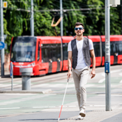 Ein junger blinder Mann läuft auf dem Gehweg. Er hat eine Sonnenbrille auf und einen Blindenstock in der Hand. Mit dem Blindenstock benutzt er die Leitstreifen auf dem Boden. Im Hintergrund fährt eine Straßenbahn entlang.