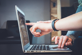 Zwei Personen arbeiten gemeinsam an einem Laptop. Eine Person zeigt mit dem Finger auf den Bildschirm, während die andere Person die Tastatur bedient. Beide Hände sind im Vordergrund zu sehen, und eine der Personen trägt ein Armband. Der Hintergrund ist unscharf, wodurch der Fokus auf den Laptop und die Hände gelenkt wird.