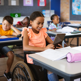 In einem Klassenzimmer sitzen mehrere Kinder an ihren Tischen und arbeiten konzentriert. Im Vordergrund sitzt ein Mädchen im Rollstuhl, das ein Tablet in den Händen hält und lächelt. Auf ihrem Tisch liegen Schulmaterialien wie Hefte und ein Federmäppchen.