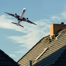 Ein Flugzeug fliegt knapp über die Dächer von Wohnhäusern. Der Himmel ist klar mit einigen Wolken, und die Dächer sind mit dunklen Ziegeln gedeckt. Ein Schornstein ist auf einem der Dächer zu sehen.