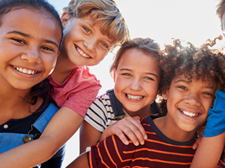 Eine Gruppe von sechs fröhlichen Kindern steht dicht beieinander und lächelt in die Kamera. Sie sind bunt gekleidet.