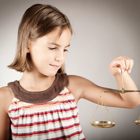 Ein junges Mädchen mit schulterlangem braunem Haar hält eine goldene Waage in ihrer rechten Hand. Sie trägt ein ärmelloses Oberteil mit Streifen in verschiedenen Farben und schaut konzentriert auf die Waage. Der Hintergrund ist neutral und grau.