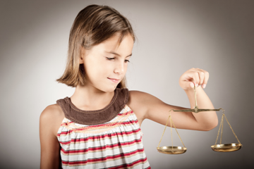 Ein junges Mädchen mit schulterlangem braunem Haar hält eine goldene Waage in ihrer rechten Hand. Sie trägt ein ärmelloses Oberteil mit Streifen in verschiedenen Farben und schaut konzentriert auf die Waage. Der Hintergrund ist neutral und grau.