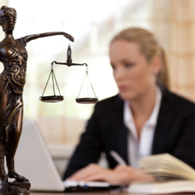 Das Bild zeigt eine Frau in einem Büro, die an einem Laptop arbeitet. Im Vordergrund steht eine Statue der Justitia, die Göttin der Gerechtigkeit, mit einer Waage in der einen und einem Schwert in der anderen Hand. Die Frau im Hintergrund ist unscharf dargestellt, während die Statue scharf fokussiert ist. Auf dem Schreibtisch vor ihr liegt ein aufgeschlagenes Buch.