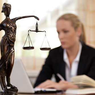 Das Bild zeigt eine Frau in einem Büro, die an einem Laptop arbeitet. Im Vordergrund steht eine Statue der Justitia, die Göttin der Gerechtigkeit, mit einer Waage in der einen und einem Schwert in der anderen Hand. Die Frau im Hintergrund ist unscharf dargestellt, während die Statue scharf fokussiert ist. Auf dem Schreibtisch vor ihr liegt ein aufgeschlagenes Buch. 
