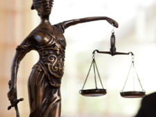 Das Bild zeigt eine Frau in einem Büro, die an einem Laptop arbeitet. Im Vordergrund steht eine Statue der Justitia, die Göttin der Gerechtigkeit, mit einer Waage in der einen und einem Schwert in der anderen Hand. Die Frau im Hintergrund ist unscharf dargestellt, während die Statue scharf fokussiert ist. Auf dem Schreibtisch vor ihr liegt ein aufgeschlagenes Buch. 