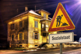 Das Bild zeigt ein altes, beleuchtetes Gebäude bei Nacht. Im Vordergrund steht ein Verkehrsschild mit einem Baustellensymbol, das einen Arbeiter zeigt, der mit einer Schaufel arbeitet. Unter dem Baustellensymbol befindet sich ein weiteres Schild mit der Aufschrift "Sozialstaat".