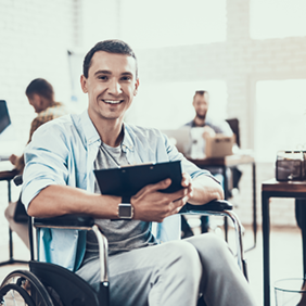 Ein Mann sitzt in einem Rollstuhl und hält ein Tablet in den Händen. Er lächelt und trägt ein hellblaues Hemd über einem grauen T-Shirt. Im Hintergrund befinden sich mehrere Personen, die an Schreibtischen arbeiten.