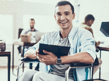 Ein Mann sitzt in einem Rollstuhl und hält ein Tablet in den Händen. Er lächelt und trägt ein hellblaues Hemd über einem grauen T-Shirt. Im Hintergrund befinden sich mehrere Personen, die an Schreibtischen arbeiten.