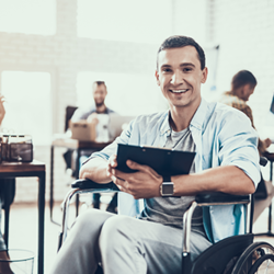 Ein Mann sitzt in einem Rollstuhl und hält ein Tablet in den Händen. Er lächelt und trägt ein hellblaues Hemd über einem grauen T-Shirt. Im Hintergrund befinden sich mehrere Personen, die an Schreibtischen arbeiten.