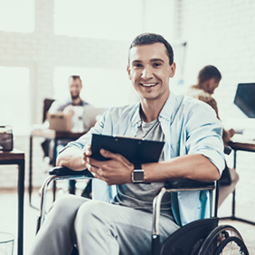 Ein Mann sitzt in einem Rollstuhl und hält ein Tablet in den Händen. Er lächelt und trägt ein hellblaues Hemd über einem grauen T-Shirt. Im Hintergrund befinden sich mehrere Personen, die an Schreibtischen arbeiten.