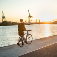 Auf dem Bild steht eine Frau mit ihrem Fahrrad an einer Kaimauer an einem Fluss. Im Hintergrund ist ein Fluss und ein Hafen zu sehen. Die Sonne geht unter.