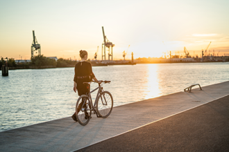 Auf dem Bild steht eine Frau mit ihrem Fahrrad an einer Kaimauer an einem Fluss. Im Hintergrund ist ein Fluss und ein Hafen zu sehen. Die Sonne geht unter.