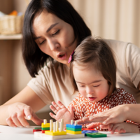 Eine Frau und ein kleines Mädchen sitzen an einem Tisch und spielen mit bunten Holzspielzeugen. Die Frau zeigt dem Mädchen, wie man die Spielzeuge benutzt, während das Mädchen konzentriert zuschaut und mit den Händen die Teile berührt. 