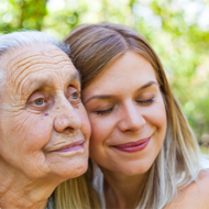 Auf dem Bild sind zwei Frauen zu sehen, die sich liebevoll aneinander schmiegen. Die jüngere Frau hat lange, blonde Haare und lächelt mit geschlossenen Augen. Die ältere Frau hat graue Haare und schaut in die Ferne. Beide befinden sich im Freien vor einem Hintergrund aus unscharfem Grün, das auf Bäume oder Pflanzen hinweist. 