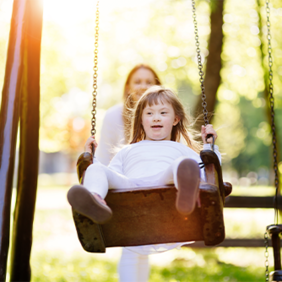 Ein Kind schaukelt auf einer Schaukel im Park. Im Hintergrund ist eine erwachsene Person zu sehen, die das Kind anschubst. Die Szene ist von hellem Sonnenlicht durchflutet, das durch die Bäume im Hintergrund scheint. Das Kind trägt helle Kleidung. 