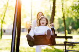 Ein Kind schaukelt auf einer Schaukel im Park. Im Hintergrund ist eine erwachsene Person zu sehen, die das Kind anschubst. Die Szene ist von hellem Sonnenlicht durchflutet, das durch die Bäume im Hintergrund scheint. Das Kind trägt helle Kleidung. 
