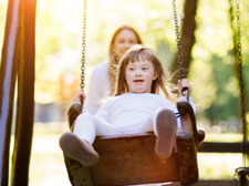 Ein Kind schaukelt auf einer Schaukel im Park. Im Hintergrund ist eine erwachsene Person zu sehen, die das Kind anschubst. Die Szene ist von hellem Sonnenlicht durchflutet, das durch die Bäume im Hintergrund scheint. Das Kind trägt helle Kleidung.