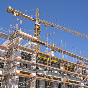 Das Bild zeigt eine Baustelle, auf der ein mehrstöckiges Gebäude errichtet wird. Das Gebäude ist von einem Gerüst umgeben, das den Arbeitern Zugang zu verschiedenen Ebenen ermöglicht. Im Hintergrund ragt ein großer Baukran in den klaren blauen Himmel, der für den Transport von Baumaterialien verwendet wird.