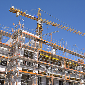 Das Bild zeigt eine Baustelle, auf der ein mehrstöckiges Gebäude errichtet wird. Das Gebäude ist von einem Gerüst umgeben, das den Arbeitern Zugang zu verschiedenen Ebenen ermöglicht. Im Hintergrund ragt ein großer Baukran in den klaren blauen Himmel, der für den Transport von Baumaterialien verwendet wird.