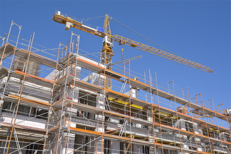Das Bild zeigt eine Baustelle, auf der ein mehrstöckiges Gebäude errichtet wird. Das Gebäude ist von einem Gerüst umgeben, das den Arbeitern Zugang zu verschiedenen Ebenen ermöglicht. Im Hintergrund ragt ein großer Baukran in den klaren blauen Himmel, der für den Transport von Baumaterialien verwendet wird.