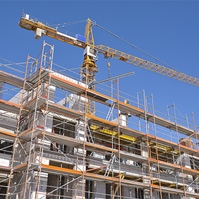 Das Bild zeigt eine Baustelle, auf der ein mehrstöckiges Gebäude errichtet wird. Das Gebäude ist von einem Gerüst umgeben, das den Arbeitern Zugang zu verschiedenen Ebenen ermöglicht. Im Hintergrund ragt ein großer Baukran in den klaren blauen Himmel, der für den Transport von Baumaterialien verwendet wird.