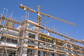 Das Bild zeigt eine Baustelle, auf der ein mehrstöckiges Gebäude errichtet wird. Das Gebäude ist von einem Gerüst umgeben, das den Arbeitern Zugang zu verschiedenen Ebenen ermöglicht. Im Hintergrund ragt ein großer Baukran in den klaren blauen Himmel, der für den Transport von Baumaterialien verwendet wird.