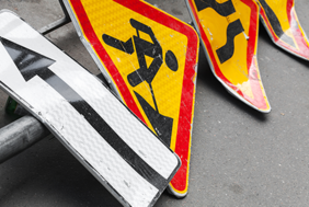 Auf dem Bild sind mehrere Verkehrsschilder zu sehen, die auf dem Boden liegen. Eines der Schilder zeigt einen schwarzen Pfeil, der nach links weist, auf weißem Hintergrund. Ein anderes Schild zeigt ein Symbol für Straßenbauarbeiten: eine stilisierte Figur, die mit einer Schaufel arbeitet, auf gelbem Hintergrund mit rotem Rand. Ein weiteres Schild zeigt eine kurvige Straße, ebenfalls auf gelbem Hintergrund mit rotem Rand.