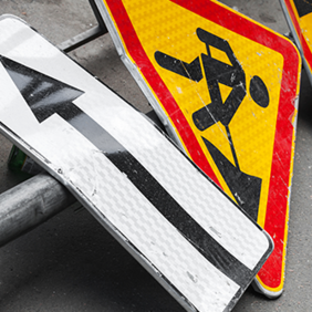 Auf dem Bild sind mehrere Verkehrsschilder zu sehen, die auf dem Boden liegen. Eines der Schilder zeigt einen schwarzen Pfeil, der nach links weist, auf weißem Hintergrund. Ein anderes Schild zeigt ein Symbol für Straßenbauarbeiten: eine stilisierte Figur, die mit einer Schaufel arbeitet, auf gelbem Hintergrund mit rotem Rand. Ein weiteres Schild zeigt eine kurvige Straße, ebenfalls auf gelbem Hintergrund mit rotem Rand.