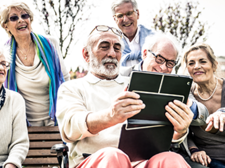 Eine Gruppe älterer Menschen sitzt und steht in einem Park. Sie schauen gemeinsam auf ein Tablet, das von einem Mann in der Mitte gehalten wird. Einige der Personen lachen. Im Hintergrund sind Bäume und ein Gebäude zu sehen.