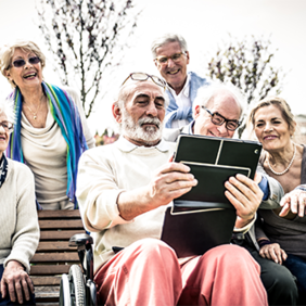 Eine Gruppe älterer Menschen sitzt und steht in einem Park. Sie schauen gemeinsam auf ein Tablet, das von einem Mann in der Mitte gehalten wird. Einige der Personen lachen. Im Hintergrund sind Bäume und ein Gebäude zu sehen.