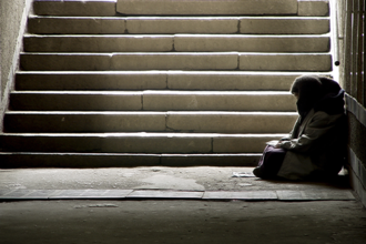 Das Bild zeigt eine Person, die in einem dunklen Durchgang oder einer Unterführung sitzt. Die Person ist in warme Kleidung gehüllt und sitzt auf dem Boden, an die Wand gelehnt. Im Hintergrund sind steinerne Treppenstufen zu sehen, die nach oben führen und von Licht durchflutet werden.