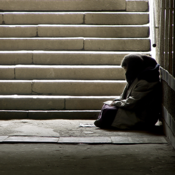 Das Bild zeigt eine Person, die in einem dunklen Durchgang oder einer Unterführung sitzt. Die Person ist in warme Kleidung gehüllt und sitzt auf dem Boden, an die Wand gelehnt. Im Hintergrund sind steinerne Treppenstufen zu sehen, die nach oben führen und von Licht durchflutet werden.