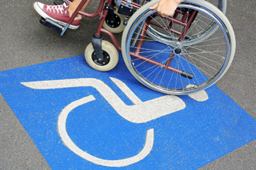 Auf dem Betonboden ist das Symbol für Rollstuhlfahrer abgebildet. Das Symbol ist in weiß auf blauen Grund. Im oberen Teil des Bildes sieht man einen Rollstuhl auf dem eine Person sitzt.