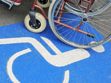 Auf dem Betonboden ist das Symbol für Rollstuhlfahrer abgebildet. Das Symbol ist in weiß auf blauen Grund. Im oberen Teil des Bildes sieht man einen Rollstuhl auf dem eine Person sitzt.
