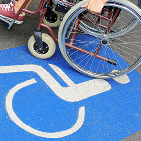 Auf dem Betonboden ist das Symbol für Rollstuhlfahrer abgebildet. Das Symbol ist in weiß auf blauen Grund. Im oberen Teil des Bildes sieht man einen Rollstuhl auf dem eine Person sitzt.