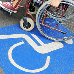 Auf dem Betonboden ist das Symbol für Rollstuhlfahrer abgebildet. Das Symbol ist in weiß auf blauen Grund. Im oberen Teil des Bildes sieht man einen Rollstuhl auf dem eine Person sitzt.