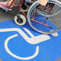 Auf dem Betonboden ist das Symbol für Rollstuhlfahrer abgebildet. Das Symbol ist in weiß auf blauen Grund. Im oberen Teil des Bildes sieht man einen Rollstuhl auf dem eine Person sitzt.