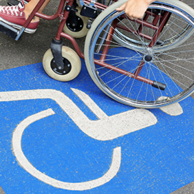 Auf dem Betonboden ist das Symbol für Rollstuhlfahrer abgebildet. Das Symbol ist in weiß auf blauen Grund. Im oberen Teil des Bildes sieht man einen Rollstuhl auf dem eine Person sitzt.