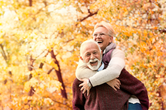 Ein älteres Paar genießt einen sonnigen Herbsttag im Freien. Die Frau, die eine Brille trägt, umarmt den Mann von hinten und beide lachen herzlich. Sie sind von herbstlich gefärbten Bäumen umgeben, deren Blätter in warmen Gelb- und Orangetönen leuchten. Der Mann trägt einen weinroten Pullover, während die Frau eine helle Jacke anhat. 