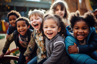 Auf dem Bild sind sechs fröhliche Kinder zu sehen, die eng beieinander stehen und in die Kamera lächeln. Sie befinden sich auf einem Spielplatz im Freien. Die Kinder tragen verschiedene farbenfrohe Kleidung. Im Hintergrund ist eine Holzkonstruktion zu erkennen.