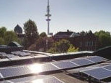 Im Vordergrund sind Solaranlagen auf einem Dach zu sehen. Im Hintergrund Bäume und in der Mitte ein Fernsehturm.