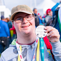 Ein junger Mann mit Brille und Baseballkappe lächelt in die Kamera und hält stolz eine Medaille hoch. Er trägt eine graue Kapuzenjacke und hat ein buntes Band um den Hals. Im Hintergrund sind weitere Personen zu sehen, die sich im Freien aufhalten. Die Atmosphäre wirkt fröhlich und feierlich.