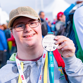 Ein junger Mann mit Brille und Baseballkappe lächelt in die Kamera und hält stolz eine Medaille hoch. Er trägt eine graue Kapuzenjacke und hat ein buntes Band um den Hals. Im Hintergrund sind weitere Personen zu sehen, die sich im Freien aufhalten. Die Atmosphäre wirkt fröhlich und feierlich.