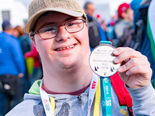 Ein junger Mann mit Brille und Baseballkappe lächelt in die Kamera und hält stolz eine Medaille hoch. Er trägt eine graue Kapuzenjacke und hat ein buntes Band um den Hals. Im Hintergrund sind weitere Personen zu sehen, die sich im Freien aufhalten. Die Atmosphäre wirkt fröhlich und feierlich.