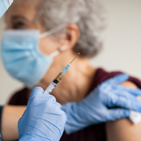 Ein medizinisches Fachpersonal, das eine blaue Maske und blaue Handschuhe trägt, verabreicht einer älteren Person eine Impfung in den Oberarm. Die ältere Person trägt ebenfalls eine Maske und ein ärmelloses Oberteil.