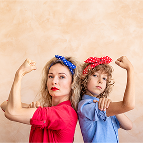 Zwei Frauen, die Rücken an Rücken stehen und ihre Arme in einer starken Pose beugen. Beide tragen bunte Haarbänder – eine Person trägt ein blaues Kopftuch mit weißen Punkten, die andere ein rotes Kopftuch mit weißen Punkten. Die Person links trägt ein rotes Hemd, während die Person rechts ein blaues Hemd trägt. Der Hintergrund ist neutral beige gehalten. Die beiden Personen schauen entschlossen in die Kamera.