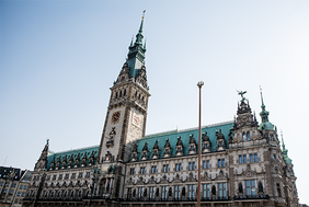 Das Bild zeigt das Hamburger Rathaus. Der Himmel im Hintergrund ist klar und blau, was das Gebäude eindrucksvoll erscheinen lässt. Das Bild ist aus der Froschperspektive gemacht.