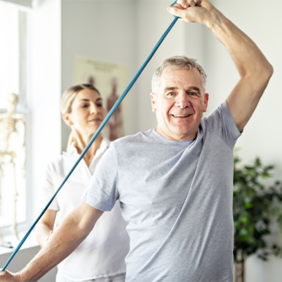 Ein älterer Mann in einem grauen T-Shirt führt eine Dehnübung mit einem blauen Widerstandsband durch. Eine Frau, möglicherweise eine Physiotherapeutin, steht hinter ihm und unterstützt ihn bei der Übung. Im Hintergrund ist ein helles Zimmer mit einem großen Fenster zu sehen, durch das Tageslicht hereinfällt.